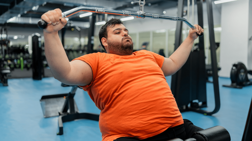 A man in an orange t-shirt performs a pulldown exercise on a machine in a gym.