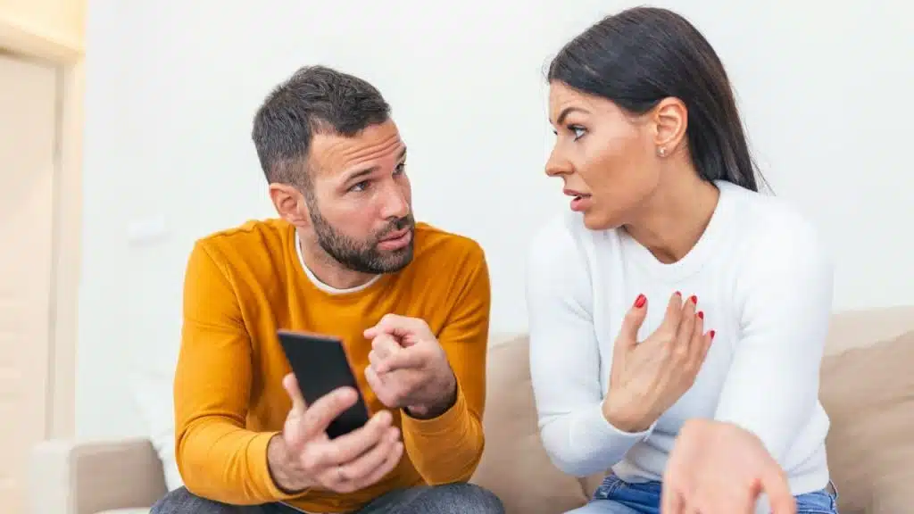 A man in a yellow shirt holds a smartphone while pointing a finger during a conversation with a woman.