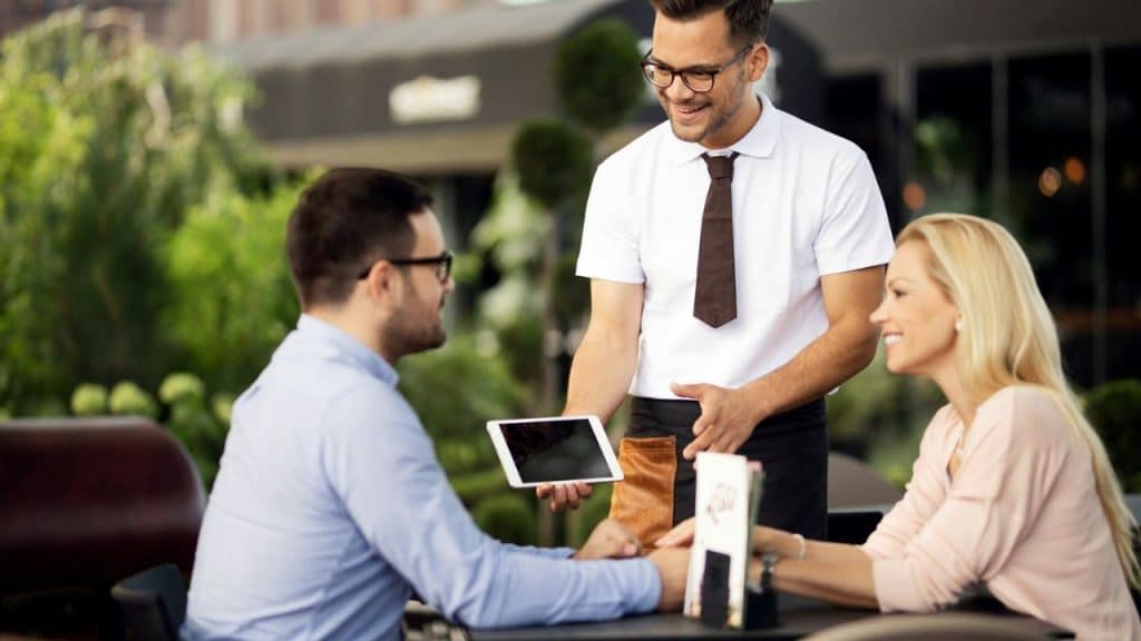 A man and woman sit at an outdoor table while a waiter holds a digital tablet.