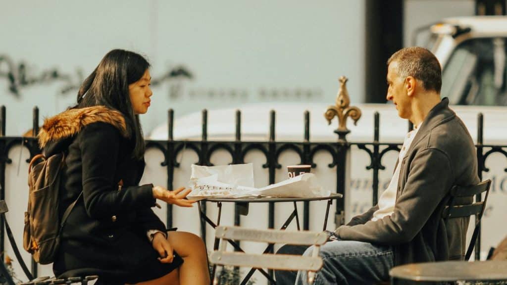 A woman and a man sit at a small outdoor table talking near a fence.