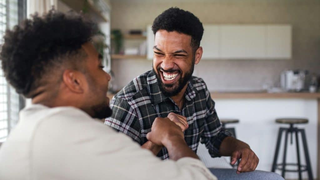 Two men bump fists and laugh while sitting together in a bright, modern indoor space.