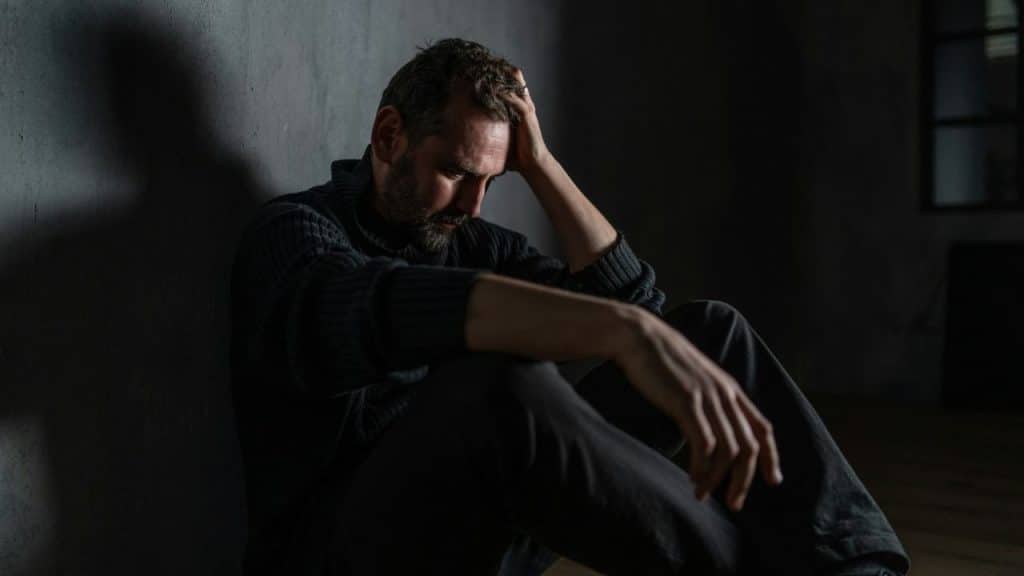 A man in a dark sweater sits on the floor against a wall, holding his head.
