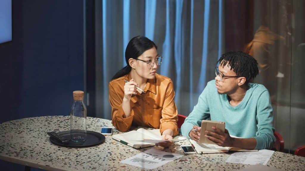 A man and a woman sit at a stone table, looking at documents and talking.
