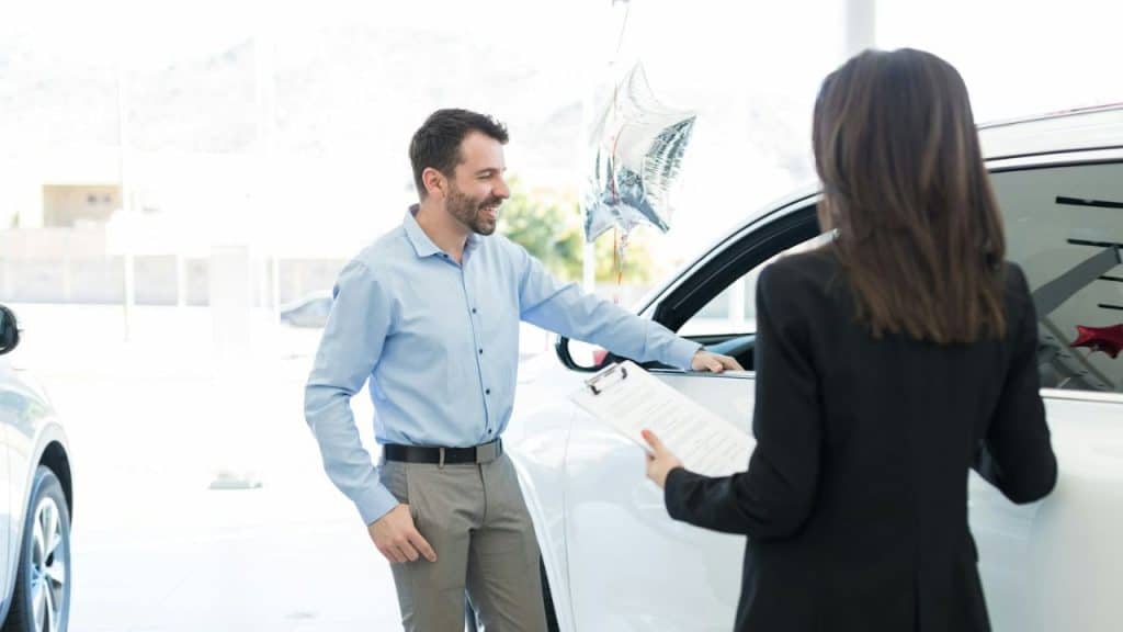 A man in a light blue shirt smiles while looking at a white car in a showroom.