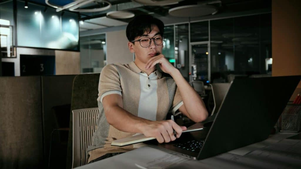A man in glasses and a polo shirt works on a laptop in an office.