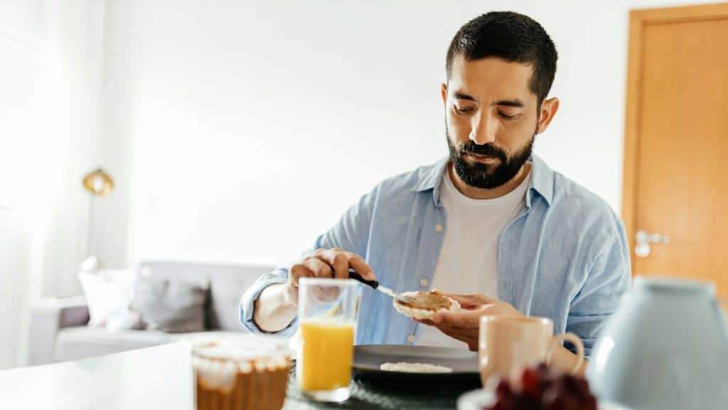 A bearded man applying butter on his toast while having breakfast at a table.