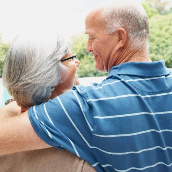 An elderly couple embracing and smiling at each other outdoors in a sunny garden setting.