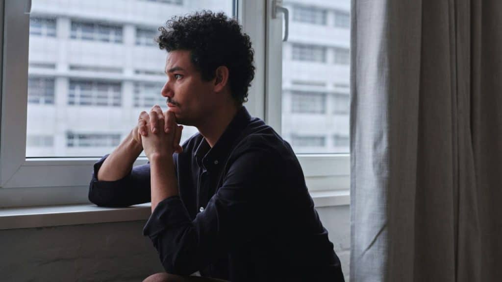 A man with curly hair sits by a window, hands clasped, looking out at a building.