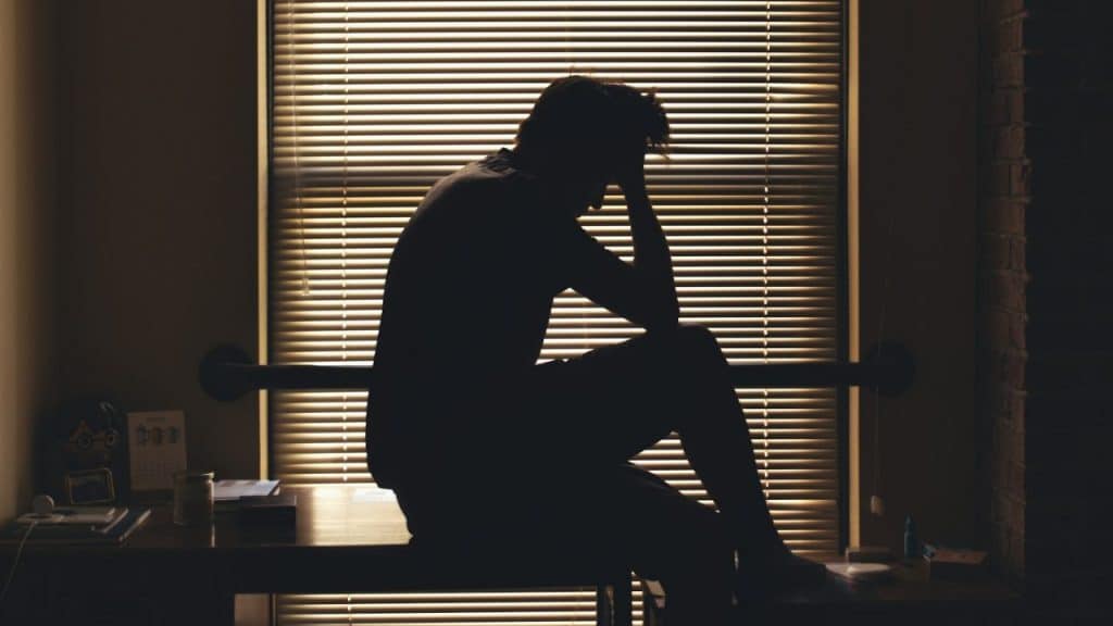 A person's dark silhouette sits hunched over on a table in front of window blinds.
