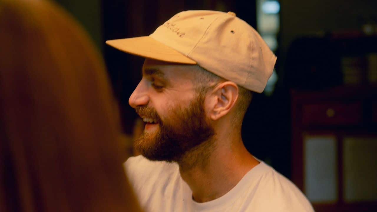 A man with a beard and a tan baseball cap smiles while looking toward another person.