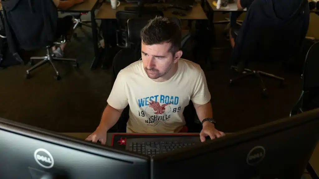 A man in a graphic t-shirt sits at a desk focusing on two large computer monitors.