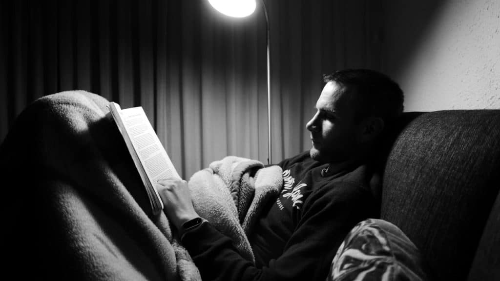 A black and white photo of a man reclining under a blanket and reading a book.