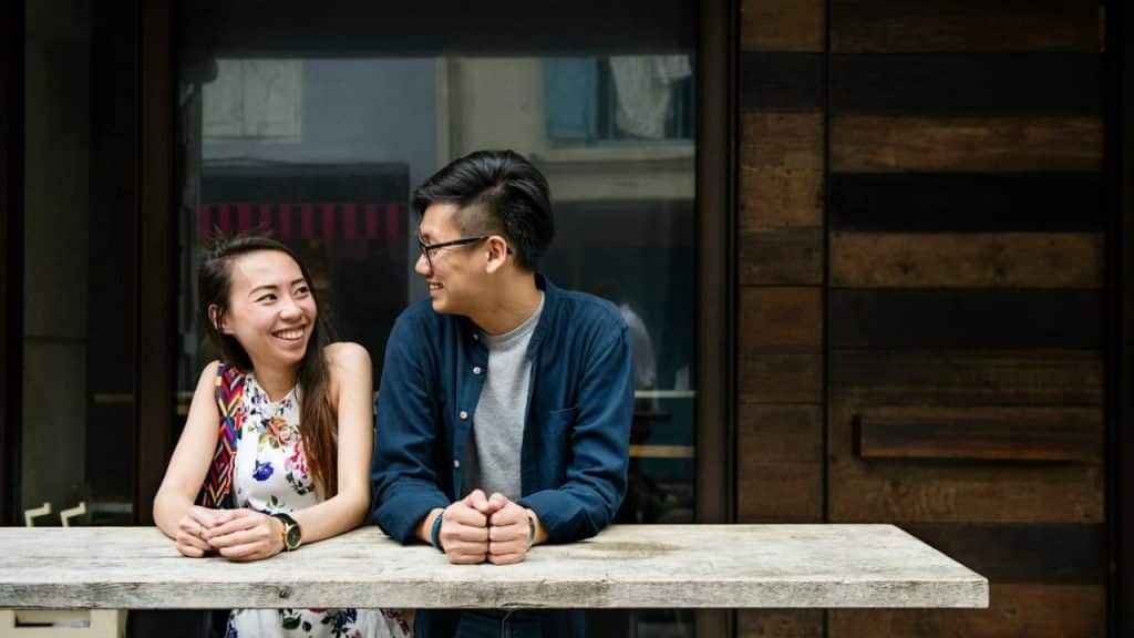 A man and woman lean on a wooden table, smiling and looking at each other.