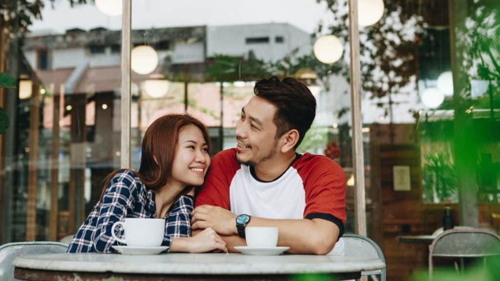 A smiling woman leans toward a man while they sit at an outdoor cafe table.