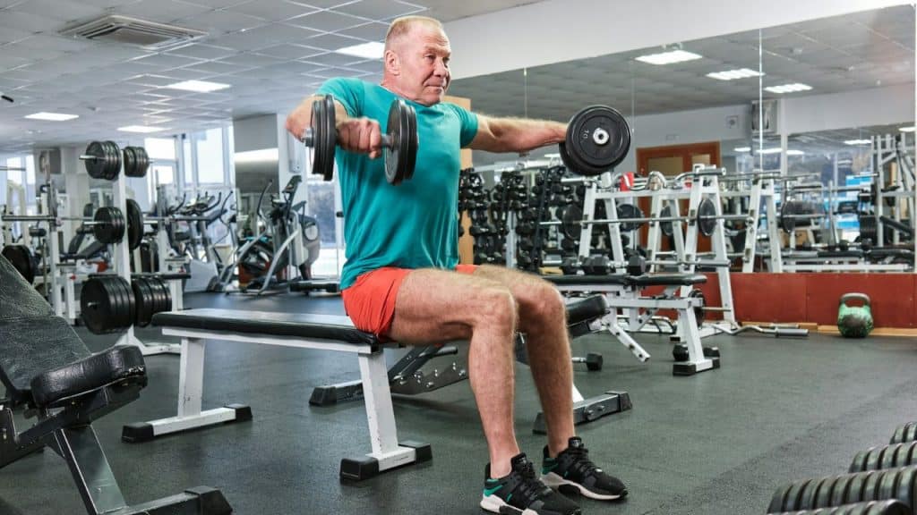 A man in a gym sitting on a bench performing lateral raises with dumbbells.