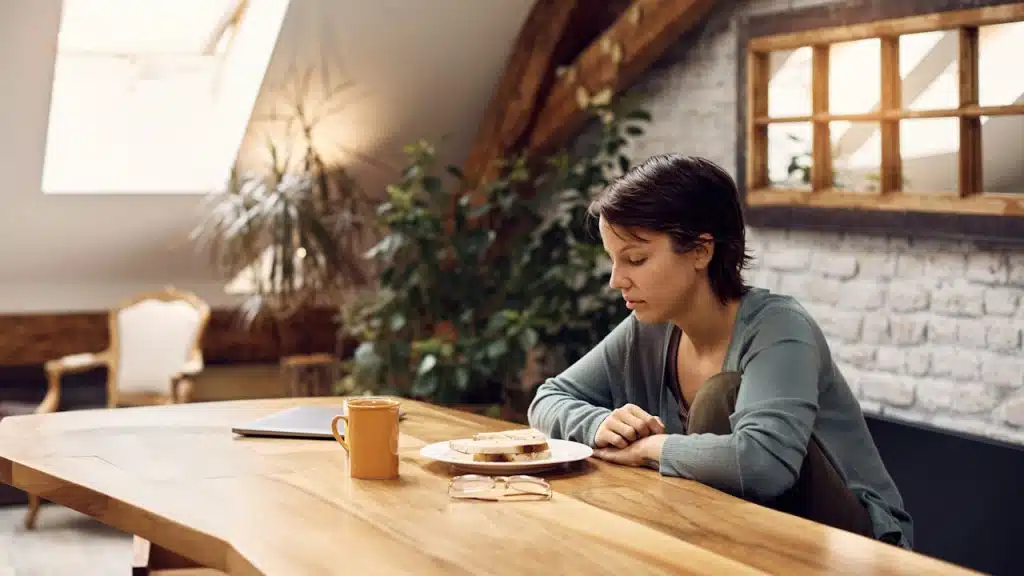A woman sits alone at a large wooden dining table in a rustic attic apartment, looking down at a plate of food with a pensive expression.