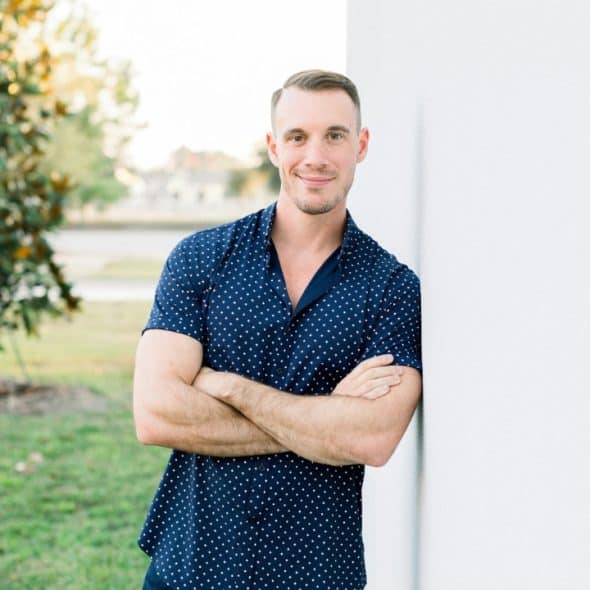 A man with short hair and a patterned shirt leans against a white wall outdoors.