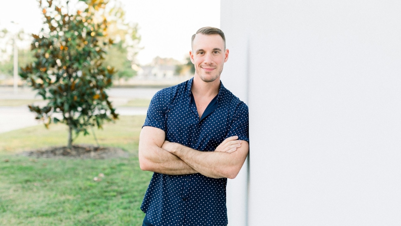 A man with short hair and a patterned shirt leans against a white wall outdoors.