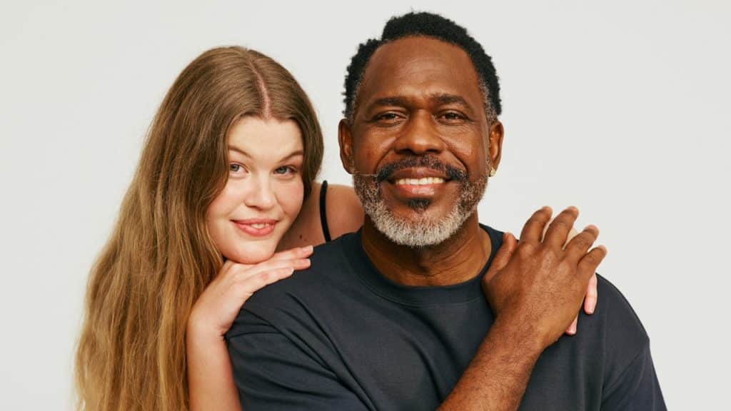 A smiling man and woman pose together against a plain white background, with the woman leaning her chin on her hands resting on the man's shoulders.