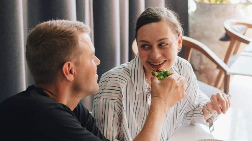 A man and woman sit at a table smiling and looking at each other while eating.