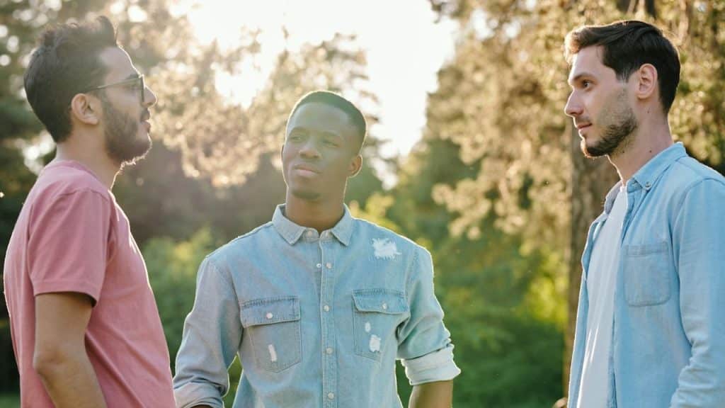 Three men stand outdoors in a sunny park setting, engaged in conversation while facing each other.