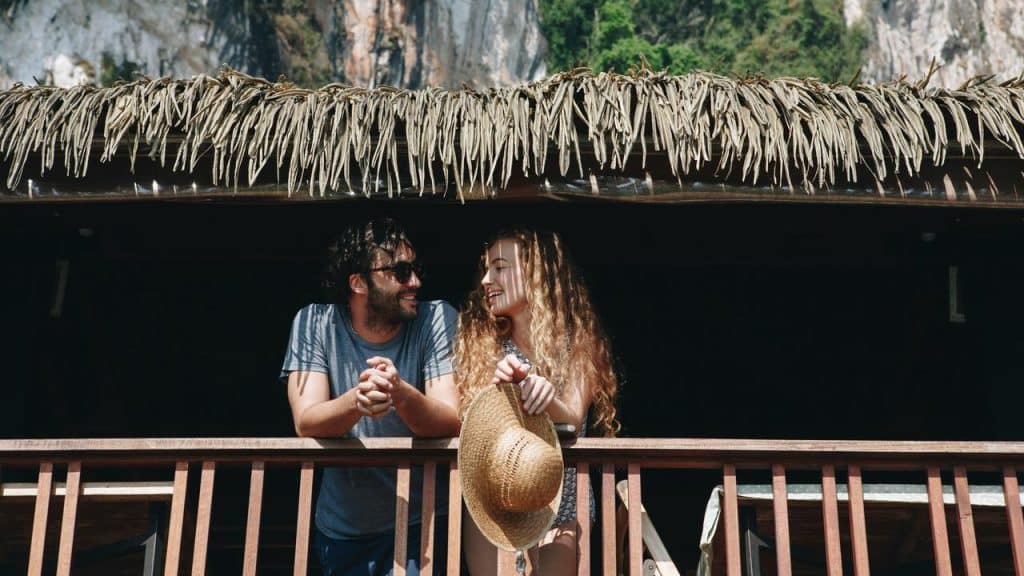 A couple chatting and smiling on a wooden balcony beneath a thatched roof.
