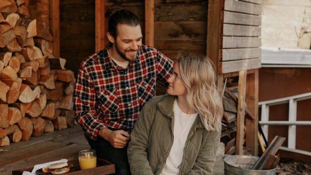 A man in a plaid shirt and a woman in a green jacket sit near a woodpile.
