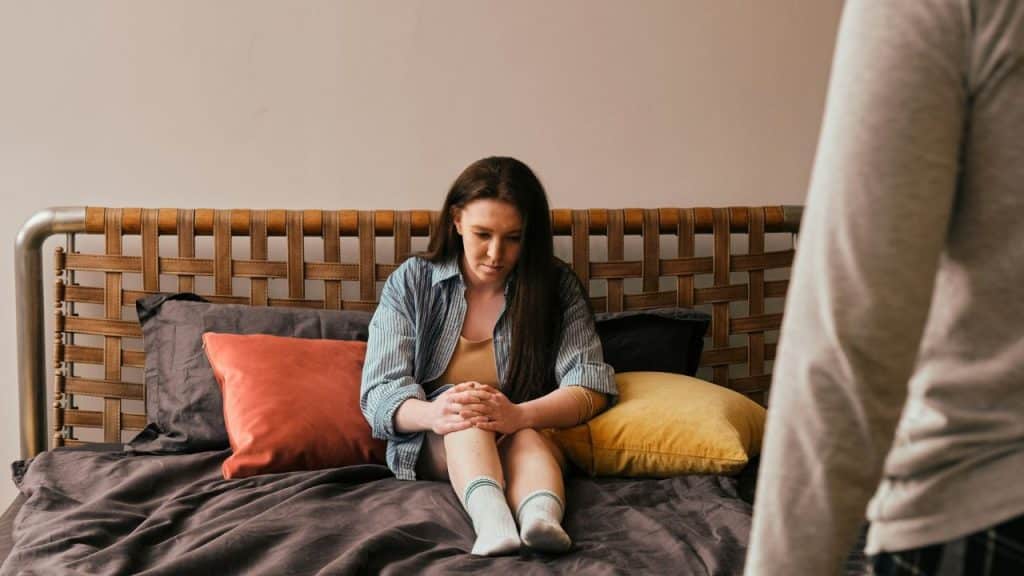 A woman sits alone on a bed looking down while someone watches from the foreground.