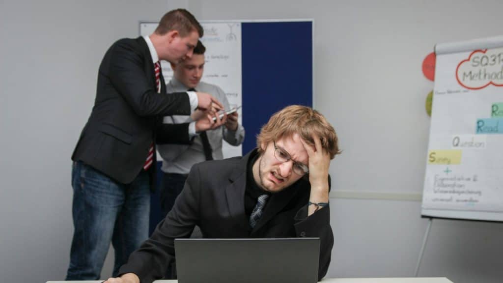 A man at a laptop holds his head while two men in suits talk behind him.