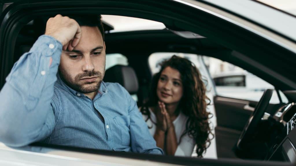 A man in a car looks down while a woman in the back seat pleads.