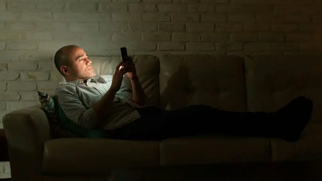 A man lies on a leather sofa in a dark room, looking at a glowing smartphone.