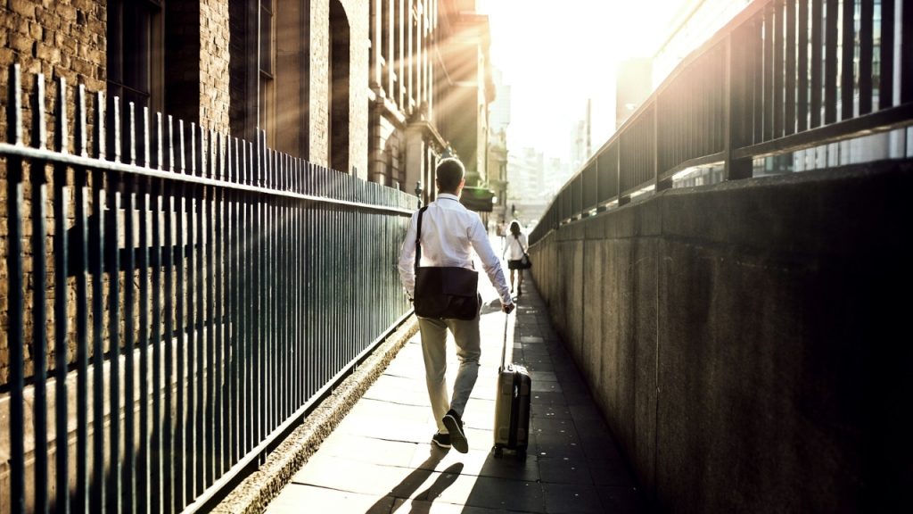 A man pulls a suitcase along a sidewalk next to a tall metal fence.
