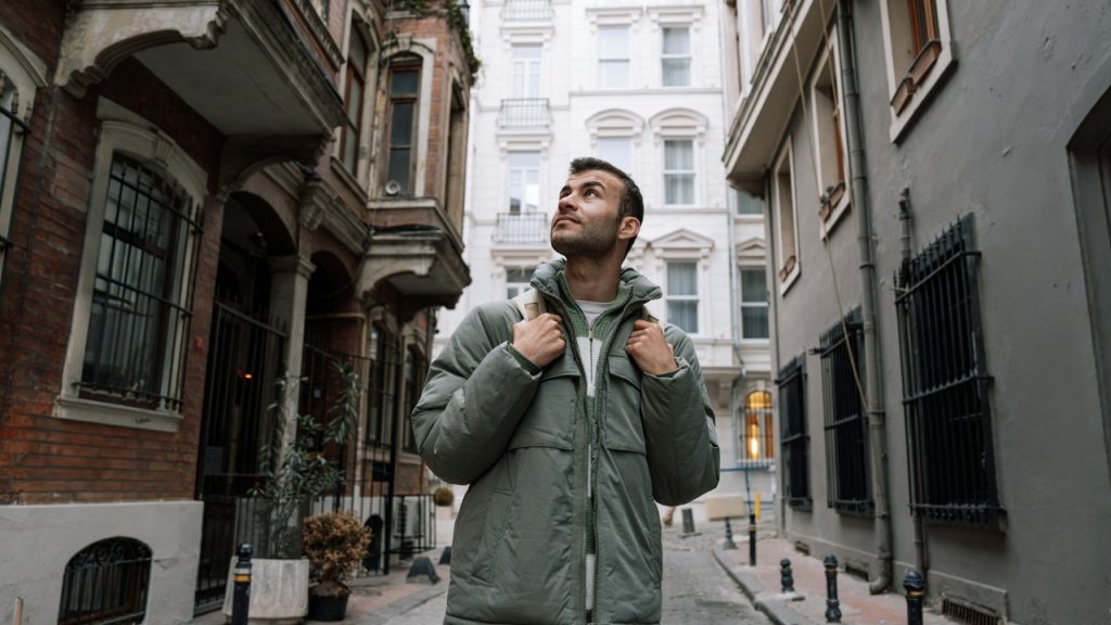 A man with a backpack stands in a narrow city street looking up at buildings.