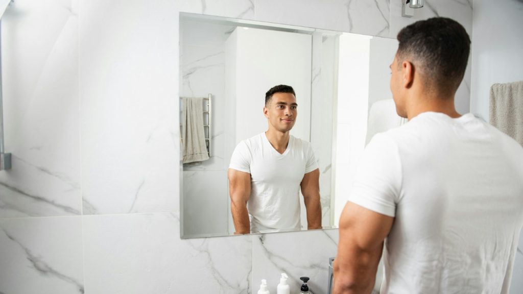 A man in a white t-shirt looks at his reflection in a bright bathroom mirror.