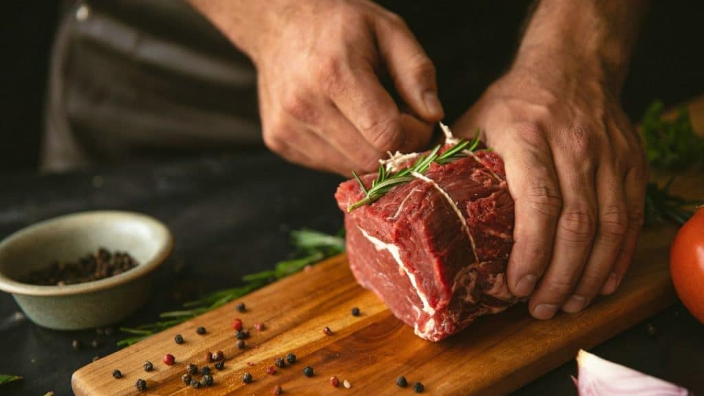 A person's hands prepare a large cut of raw red meat tied with string on a wooden board.