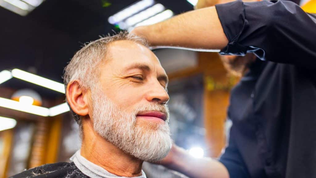 A barber styles the hair of a middle-aged man with a grey beard in a barbershop.