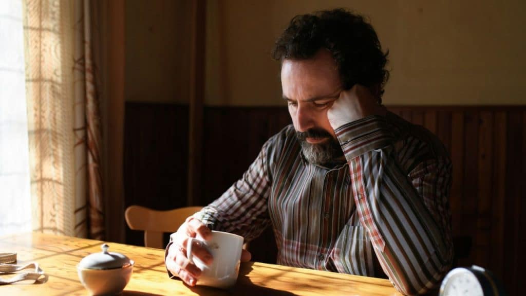 A bearded man in a striped shirt sits at a sunlit table holding a mug.