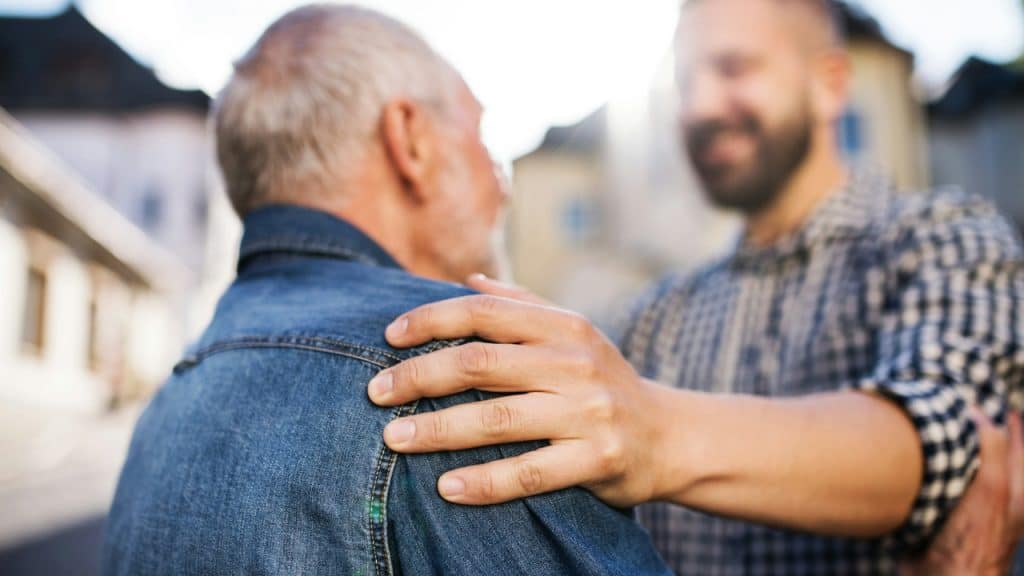 A younger man places a hand on an older man's shoulder outdoors in the sun.