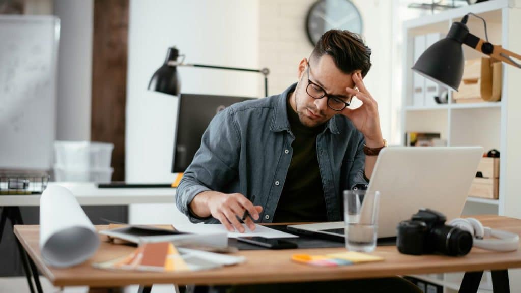 A man in glasses looks stressed while working at a desk with a laptop and papers.