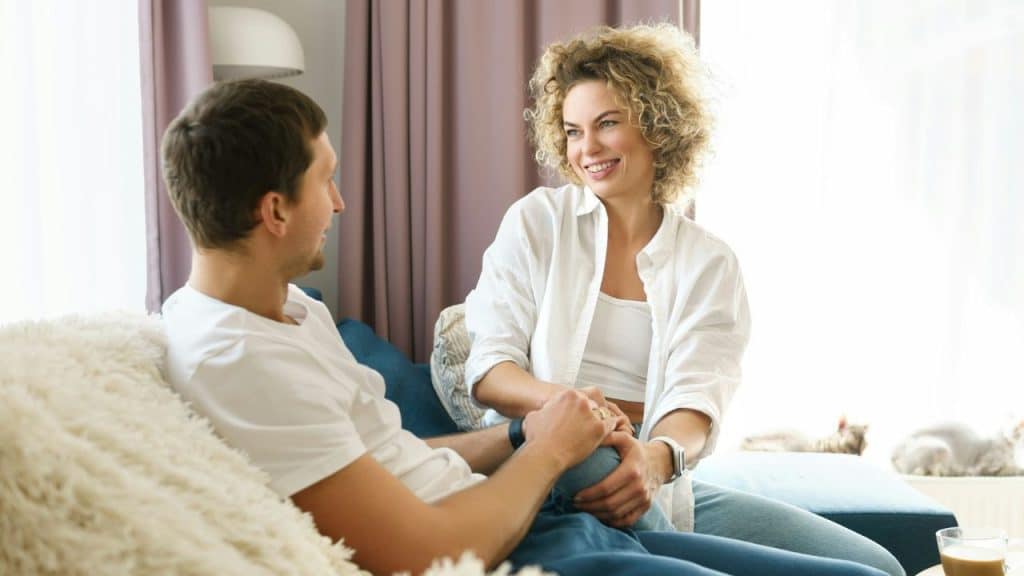 A woman with curly hair smiles while holding a man's hand on a couch.