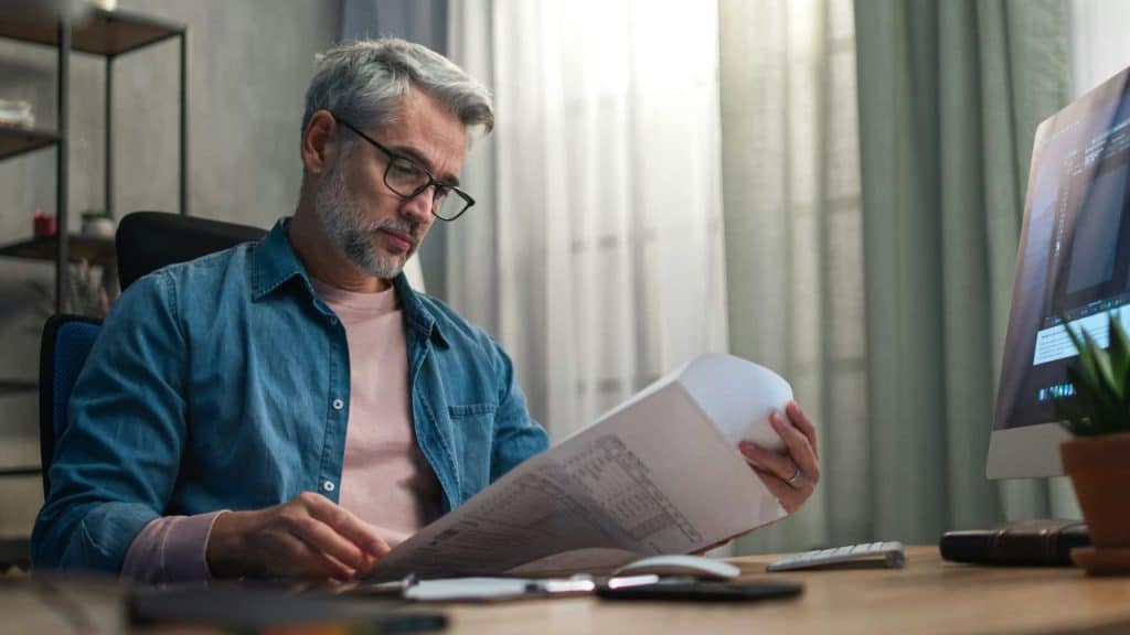 A man with gray hair and glasses sits at a desk reviewing a large document.