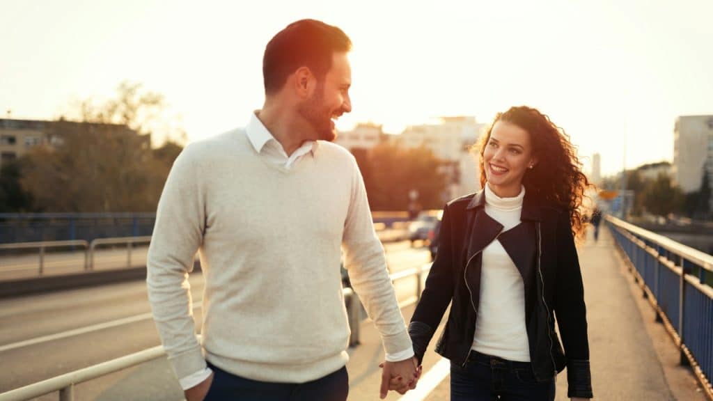 A man and woman walk across a bridge holding hands and smiling during a sunset.