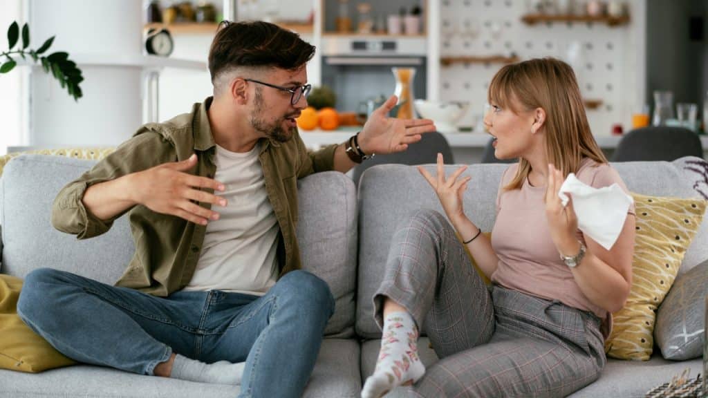 A man and woman sit on a grey sofa and gesture intensely while having a heated conversation.
