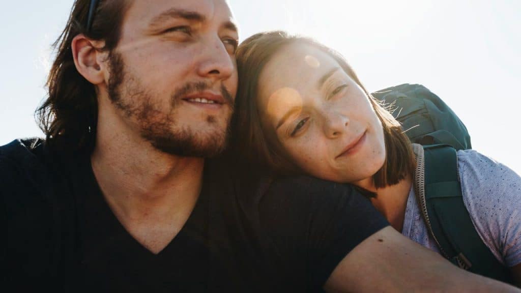 A couple leaning together outdoors, smiling softly in bright sunlight.