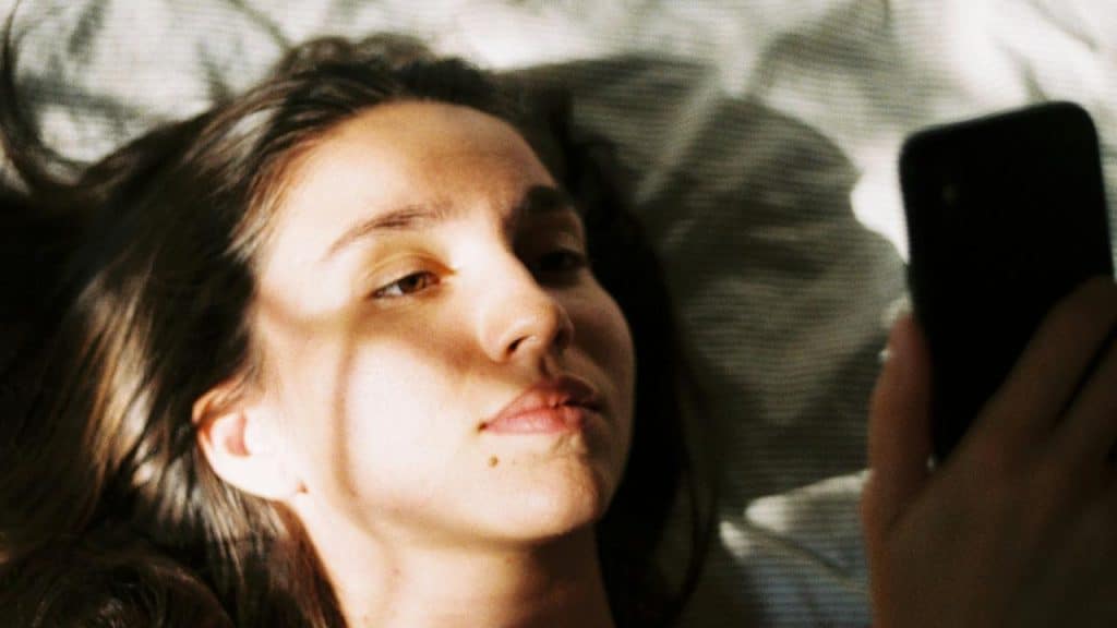 A person lying down while looking at a phone in soft, dappled light.