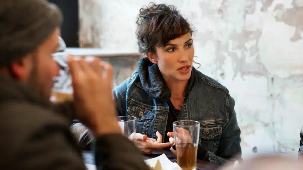 A woman talks at a table while holding a glass in a café.