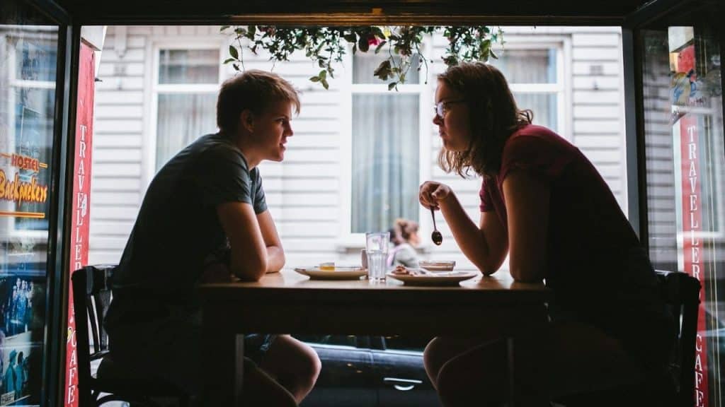 Two people sit at a small table inside a cafe, facing each other in silhouette.