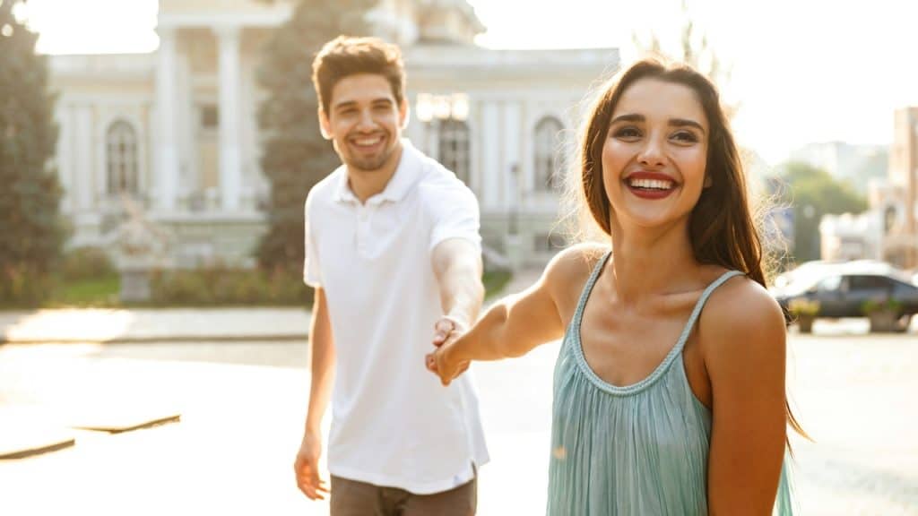 A smiling woman in a blue dress leads a man by the hand outdoors.