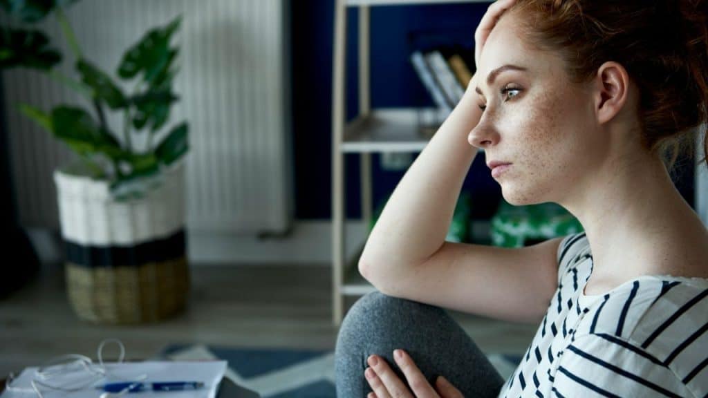 A pensive woman with freckles and red hair rests her head on her hand.