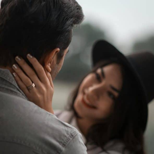 A woman in a hat gently holding a man’s face outdoors.